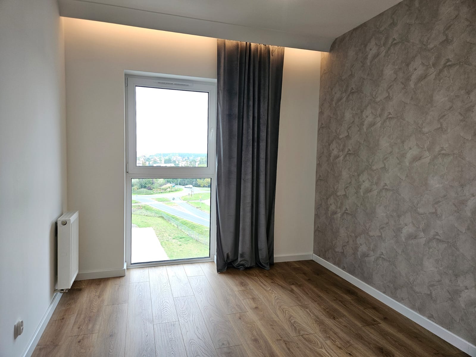 Pusty pokój po remoncie z panelami podłogowymi, ścianą z efektem dekoracyjnym, zasłoną w oknie i grzejnikiem.