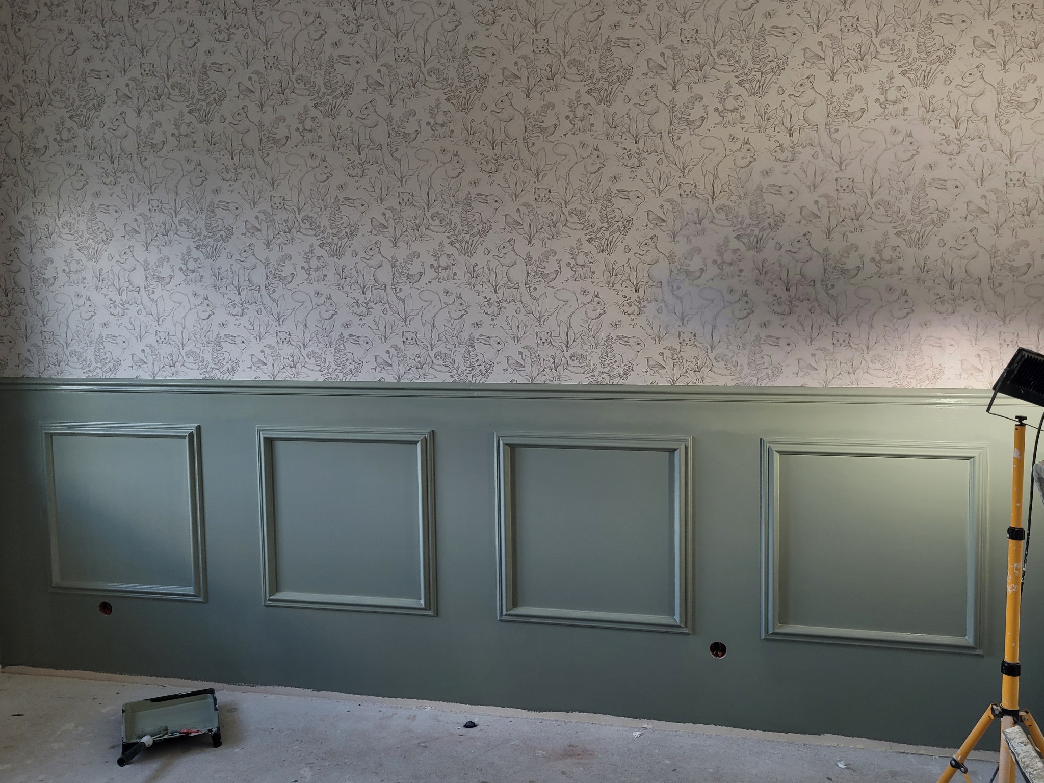 Ściana po remoncie z pomalowaną lamperią w kolorze pistacjowym i tapetą z motywem leśnych zwierząt, widoczna kuweta malarska i lampa halogenowa.