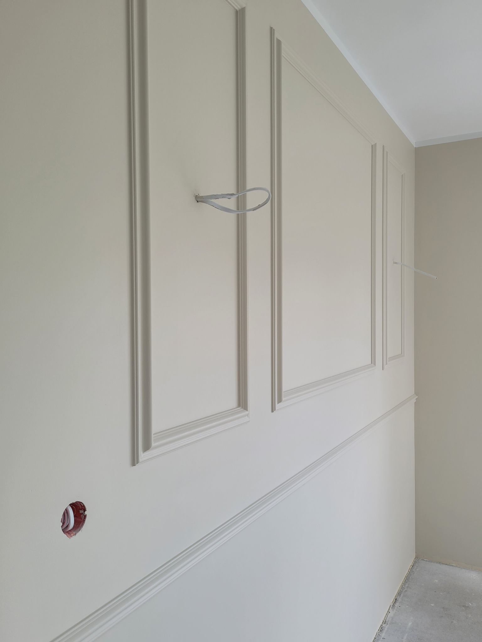 Ściana z dekoracyjnymi panelami w kolorze kremowym, widoczne przewody elektryczne wystające ze ściany i otwór na gniazdko elektryczne, surowa podłoga.