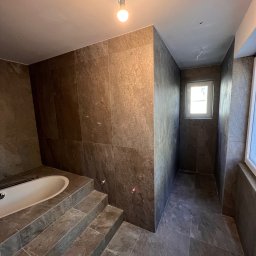 Solid Builders - Łazienka w trakcie remontu z brązowymi płytkami imitującymi kamień, widoczne schodki prowadzące do wanny, surowy strop z wiszącą żarówką i wystającymi kablami, okno w głębi pomieszczenia.