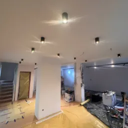 Wnętrze w trakcie remontu: widoczne punktowe oświetlenie sufitowe, zabezpieczona folią podłoga, ściany przygotowane pod gładzie, widoczne przewody elektryczne i narzędzia pracy.