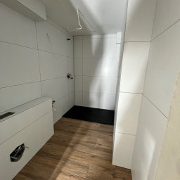 Solid Builders - Nowoczesna łazienka w trakcie wykańczania: drewnopodobna podłoga, białe płytki na ścianach, czarny brodzik, wystające kable elektryczne, otwór na rury w zabudowie geberit.