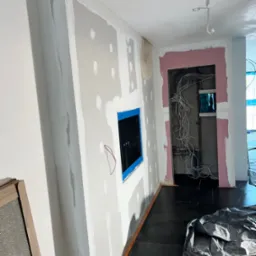 Wnętrze w trakcie remontu: ściany z nałożoną gładzią szpachlową, otwór na rozdzielnicę elektryczną zabezpieczony taśmą, widoczne przewody elektryczne w ścianie, podłoga zabezpieczona folią malarską.