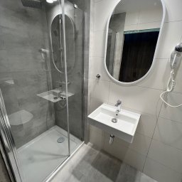 Solid Builders - Nowoczesna łazienka z kabiną prysznicową typu walk-in, umywalką na ścianie, owalnym lustrem i suszarką do włosów. Widoczny minimalistyczny design i szara kolorystyka płytek.