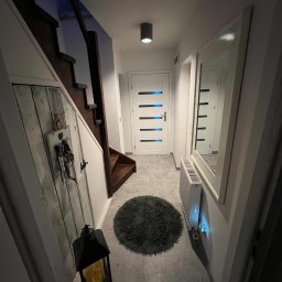 Solid Builders - Wnętrze przedpokoju z drewnianymi schodami, szarym dywanikiem, grzejnikiem, lustrem w białej ramie i lampionem ze świecami, oświetlone punktowym światłem.