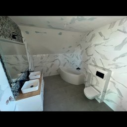 Solid Builders - Wykończona łazienka na poddaszu z marmurowymi płytkami na ścianach, szarą podłogą, białą wanną narożną, wiszącą toaletą i podwójną umywalką na drewnianym blacie.