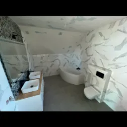 Wykończona łazienka na poddaszu z marmurowymi płytkami na ścianach, szarą podłogą, białą wanną narożną, wiszącą toaletą i podwójną umywalką na drewnianym blacie.