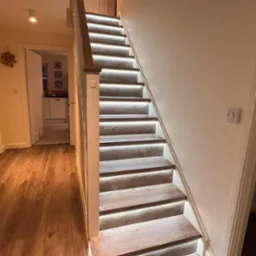 Oświetlone schody z drewnianymi stopniami i podświetleniem LED, widoczne drzwi i podłoga z paneli, ciepłe światło w pomieszczeniu.