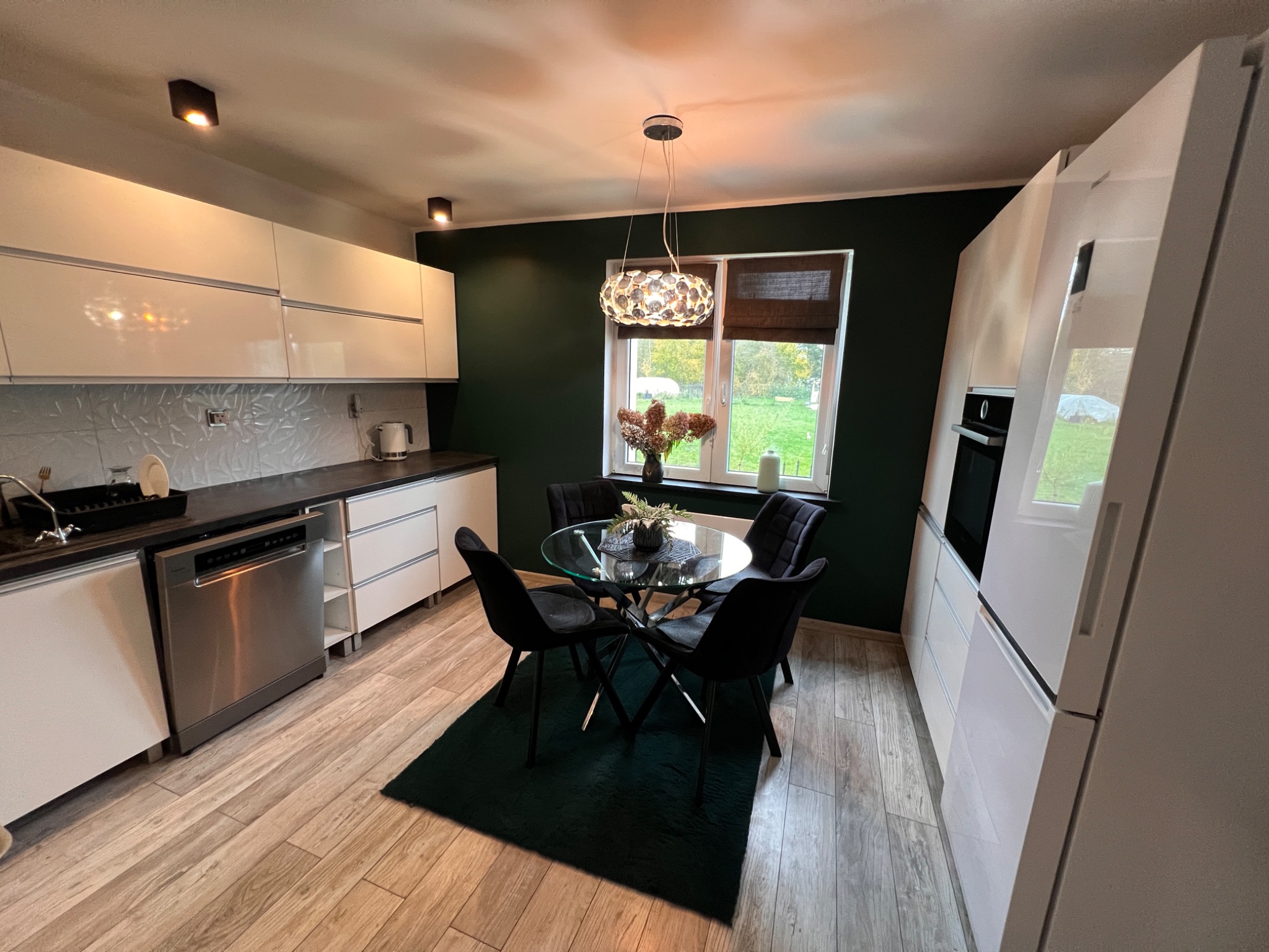 Nowoczesna kuchnia z białymi szafkami, drewnianą podłogą i ciemnozieloną ścianą, okrągły szklany stół z czarnymi krzesłami na ciemnozielonym dywanie, ozdobna lampa wisząca nad stołem.