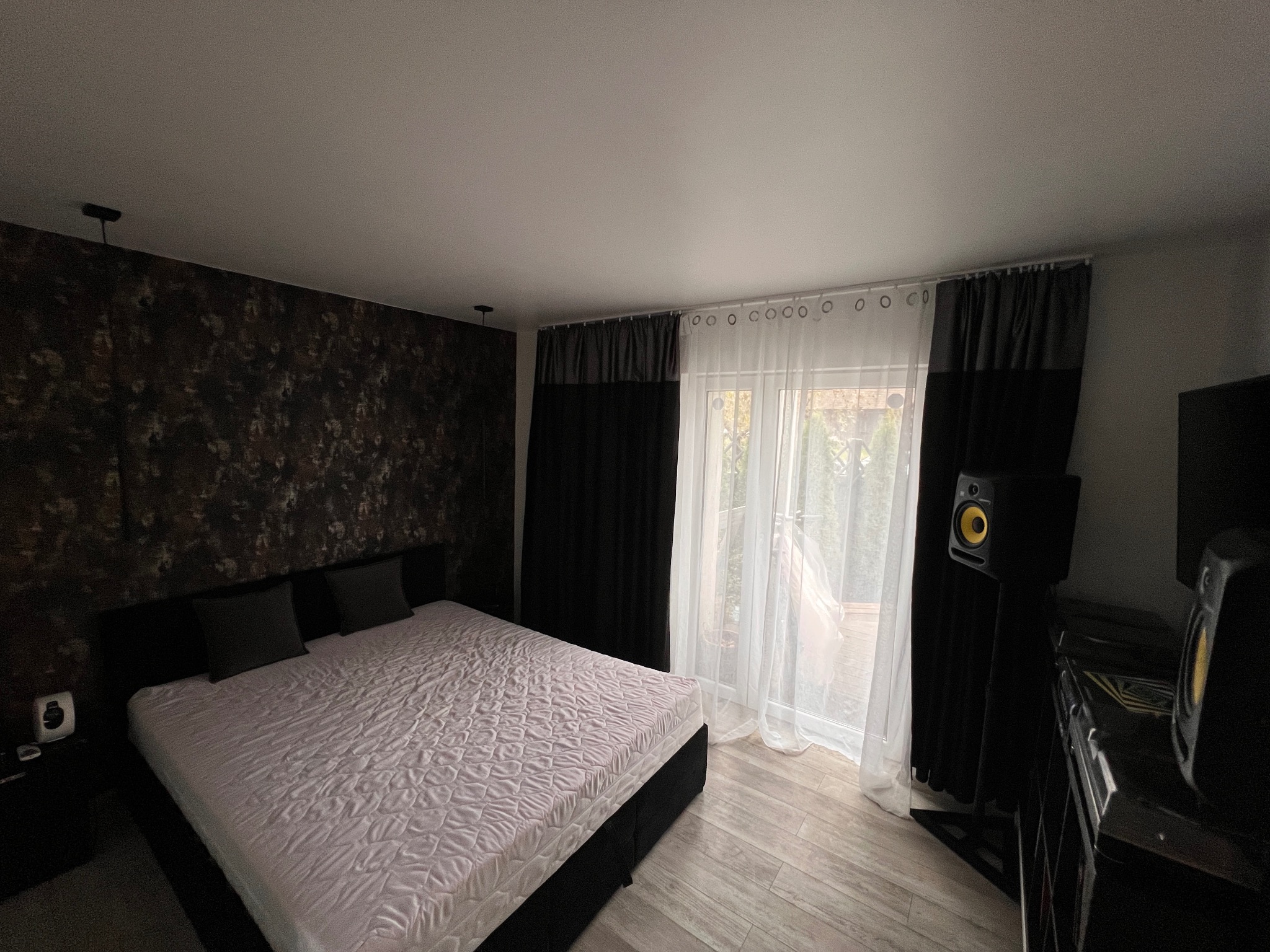 Sypialnia z łóżkiem małżeńskim, wzorzystą tapetą, czarnymi zasłonami i białymi firankami, z widocznymi głośnikami studyjnymi po prawej stronie.