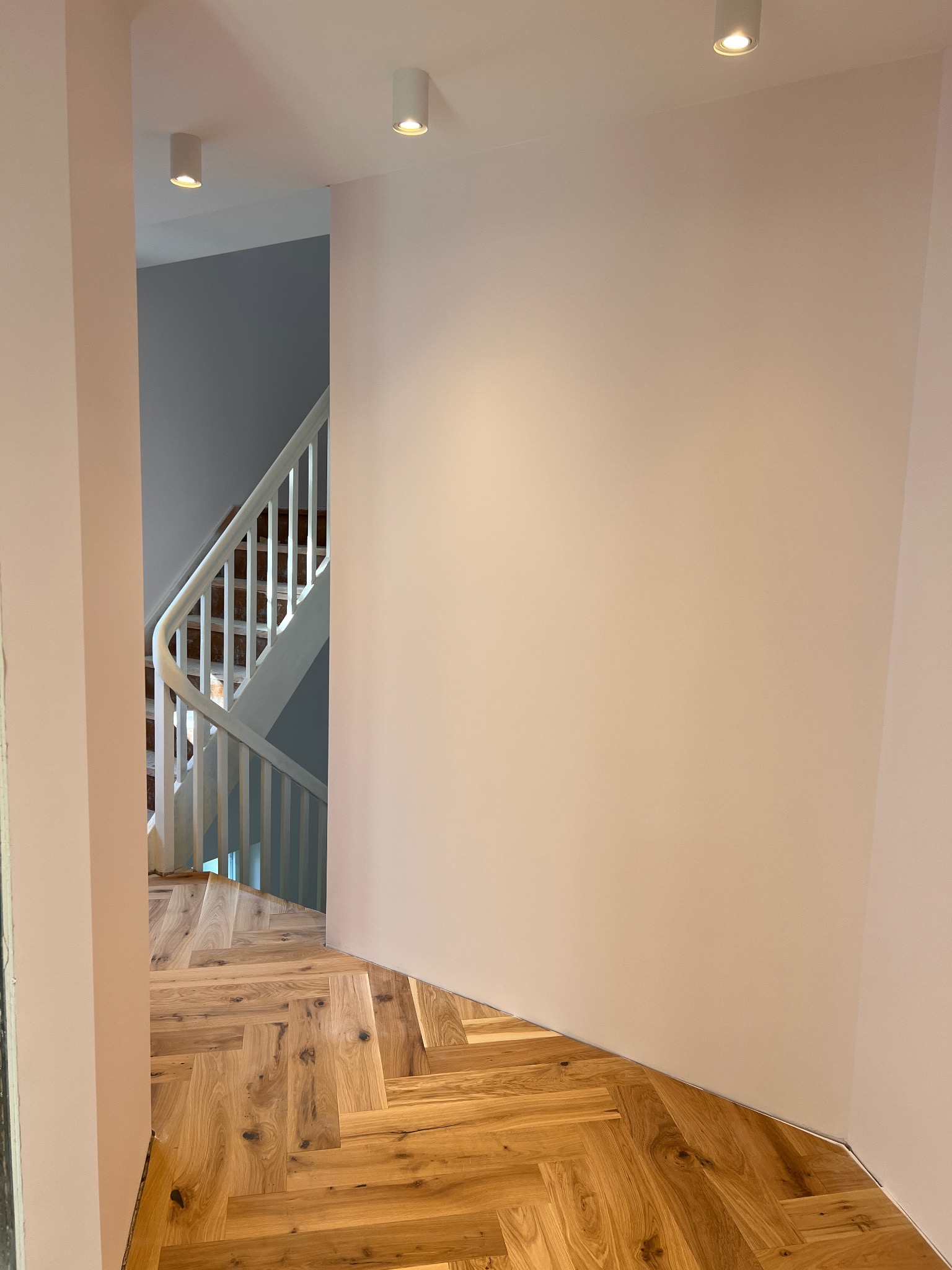 Nowo ułożony parkiet w jodełkę, świeżo pomalowane ściany na jasny róż, widok na białe schody w tle, zamontowane białe reflektory sufitowe.