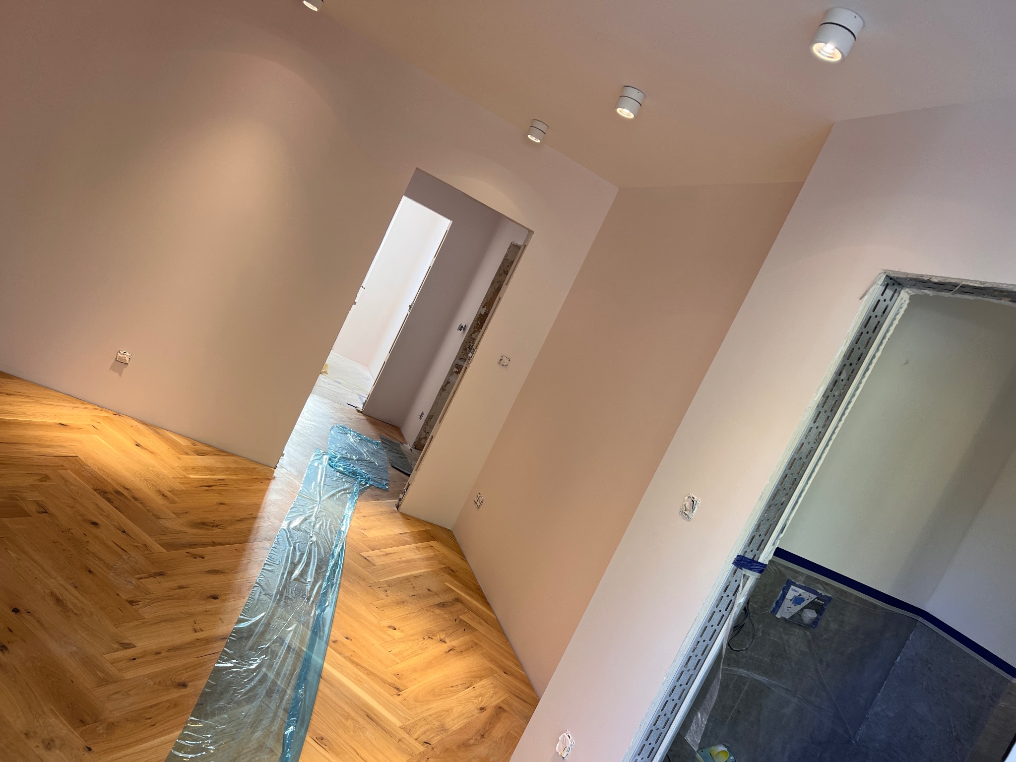 Świeżo wykończone wnętrze w Modlimowie z parkietem ułożonym w jodełkę, zabezpieczonym folią malarską, ściany pomalowane na jasny kolor, widoczne punkty oświetleniowe na suficie i przygotowane...