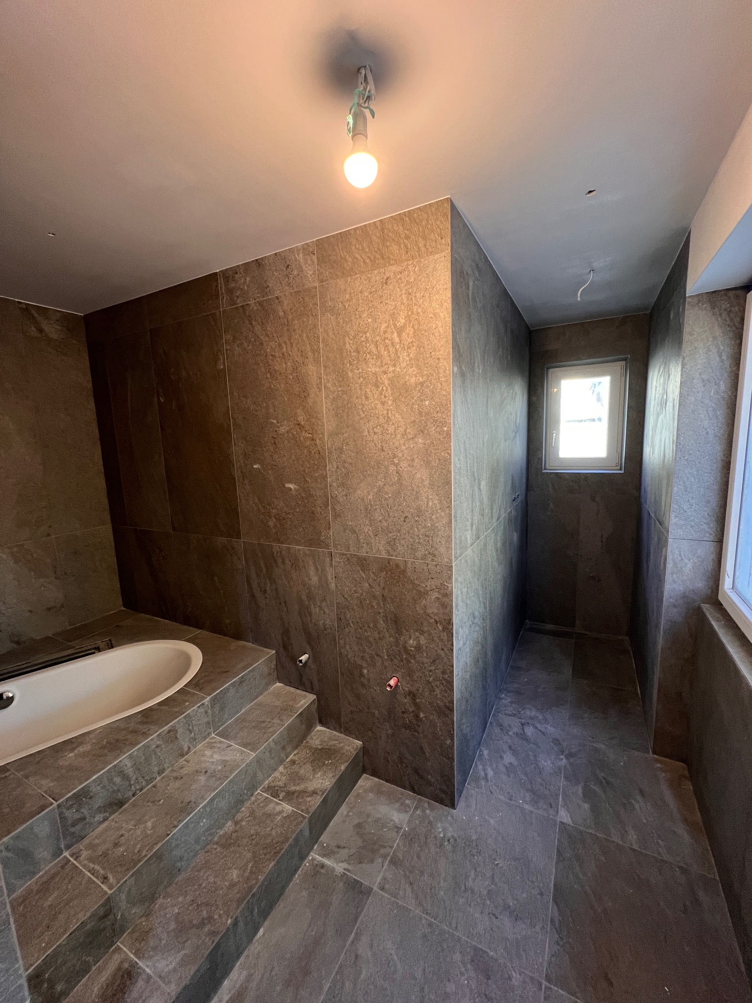 Łazienka w trakcie remontu z brązowymi płytkami imitującymi kamień, widoczne schodki prowadzące do wanny, surowy strop z wiszącą żarówką i wystającymi kablami, okno w głębi pomieszczenia.