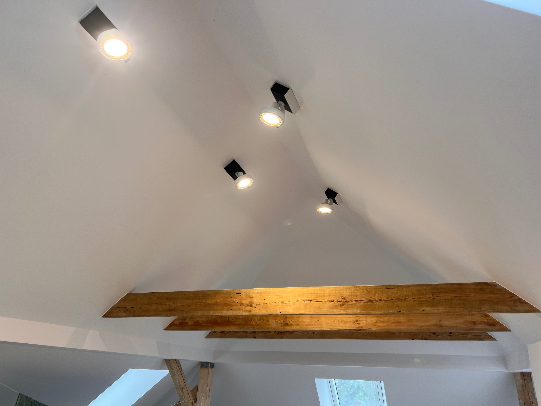 Biały sufit pod skosem z drewnianymi belkami i zamontowanymi reflektorami punktowymi z czarnymi obudowami.