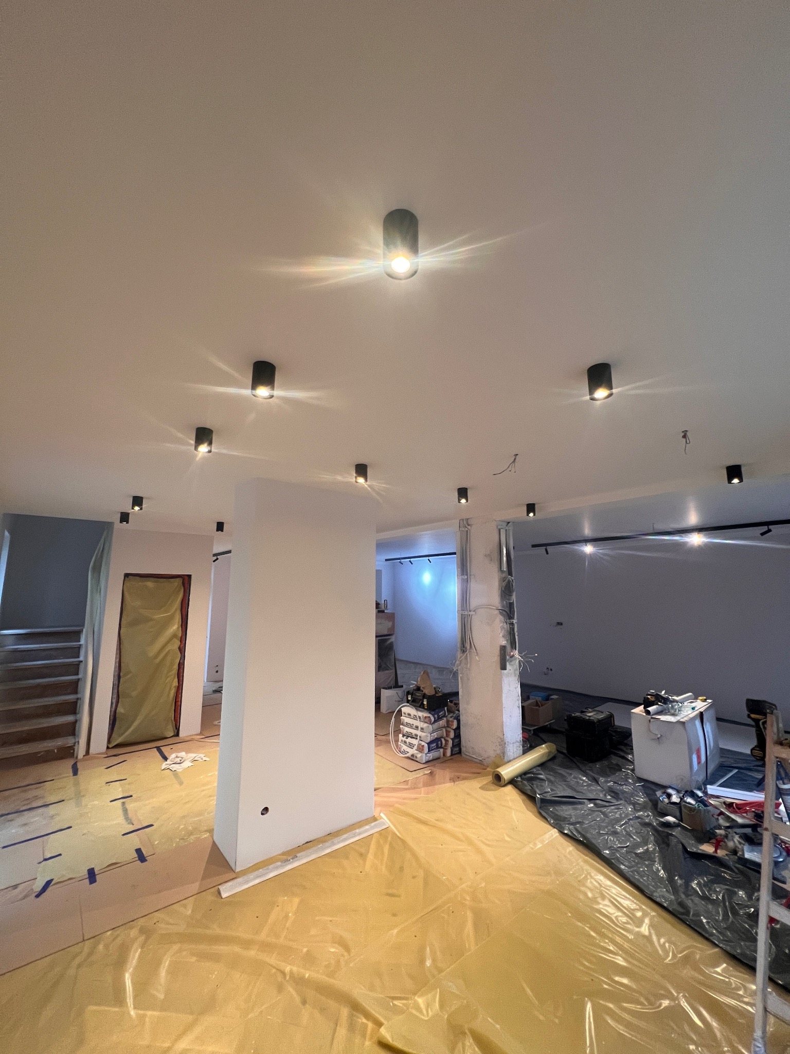 Wnętrze w trakcie remontu: widoczne punktowe oświetlenie sufitowe, zabezpieczona folią podłoga, ściany przygotowane pod gładzie, widoczne przewody elektryczne i narzędzia pracy.