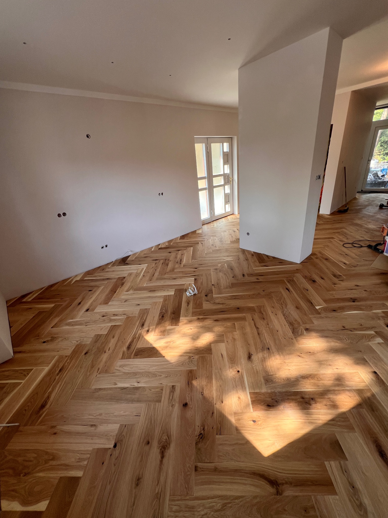 Ułożony parkiet w jodełkę w jasnym, nowym wnętrzu. Świeże, surowe ściany z widocznymi puszkami elektrycznymi. Promienie słoneczne podkreślają strukturę drewna.