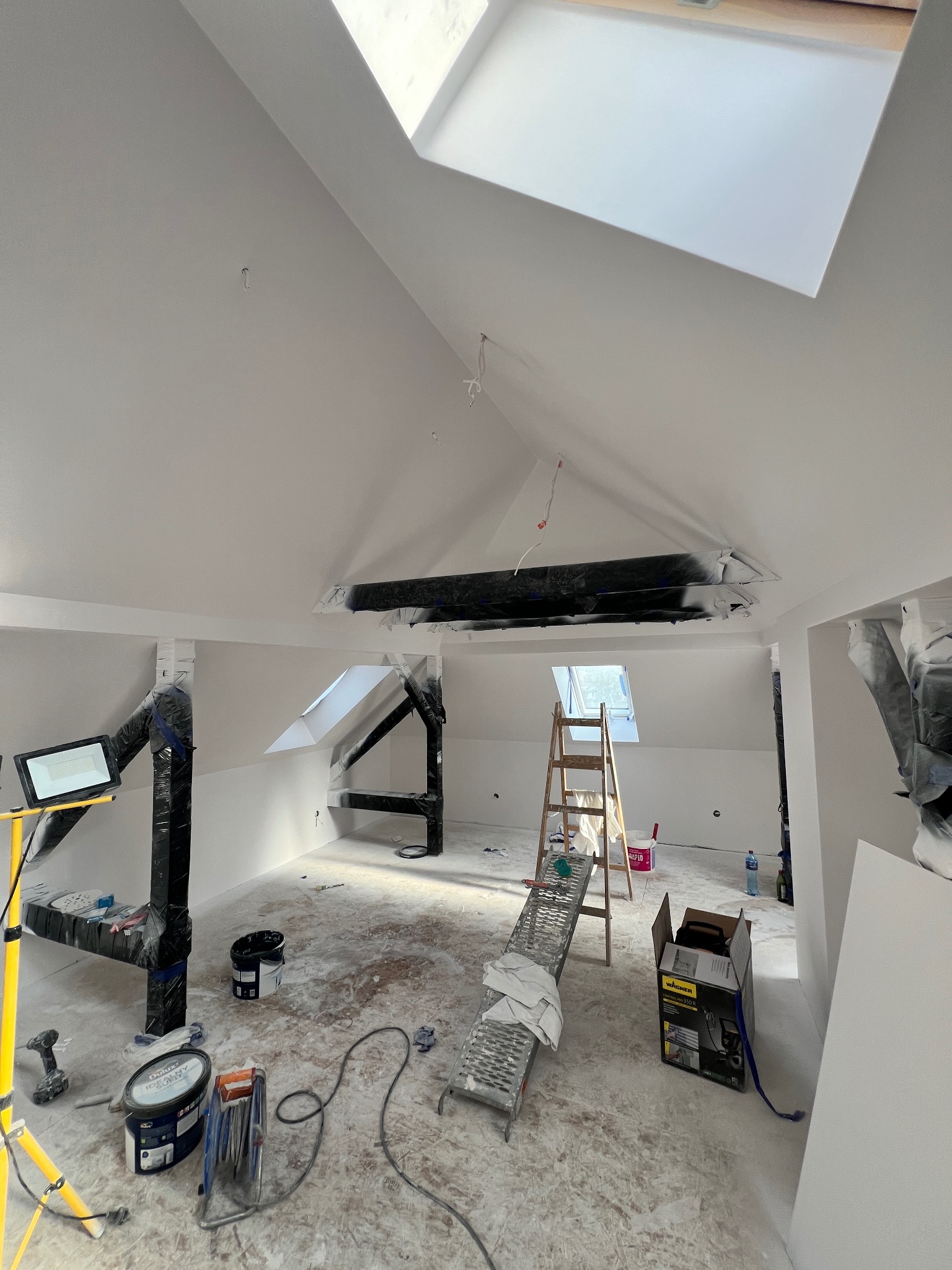 Poddasze w trakcie remontu, widoczne rusztowania zabezpieczone folią, drabina, rozłożone narzędzia malarskie, ślady farby na podłodze, okna dachowe wpuszczające dużo światła.