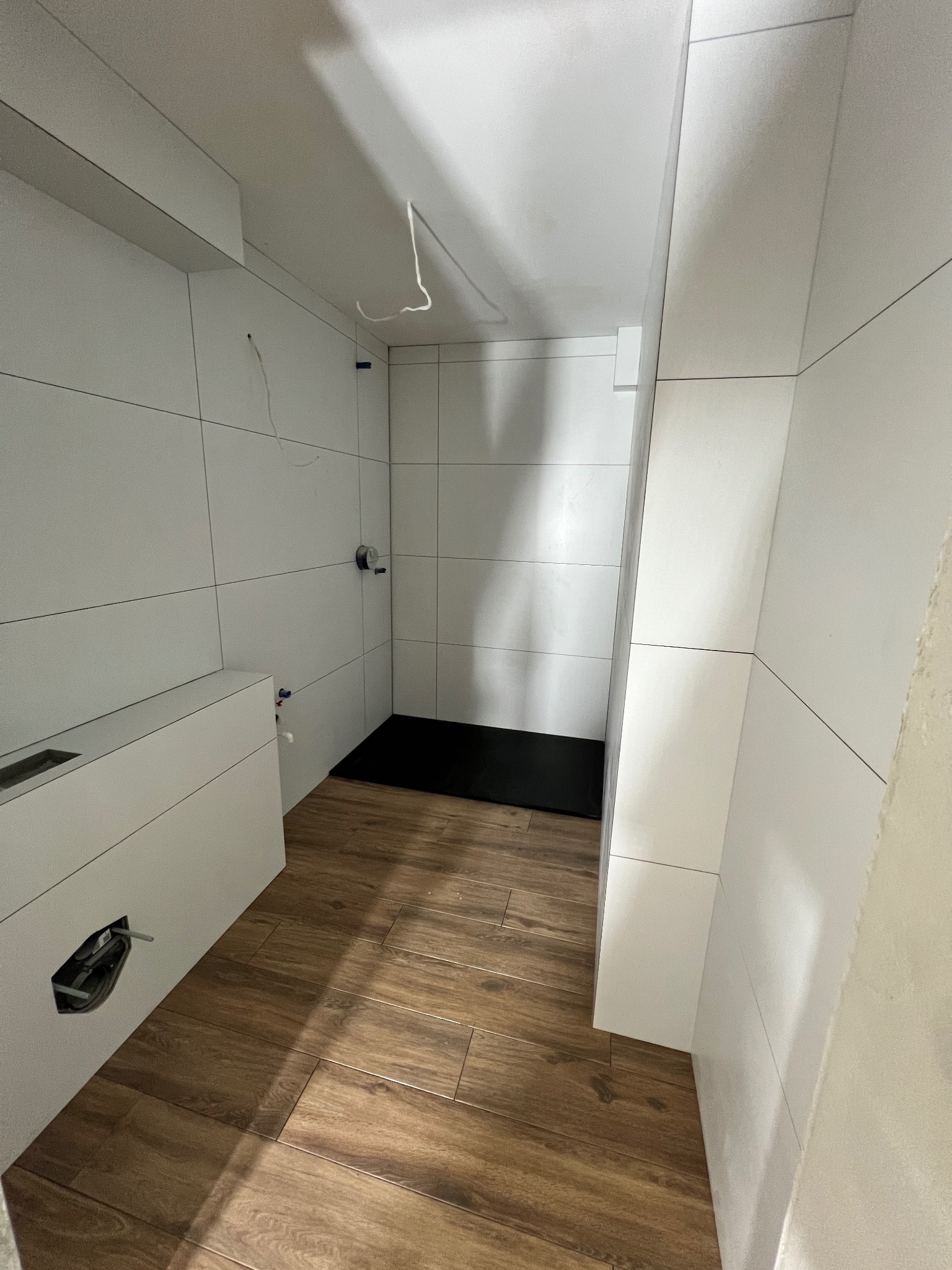 Nowoczesna łazienka w trakcie wykańczania: drewnopodobna podłoga, białe płytki na ścianach, czarny brodzik, wystające kable elektryczne, otwór na rury w zabudowie geberit.