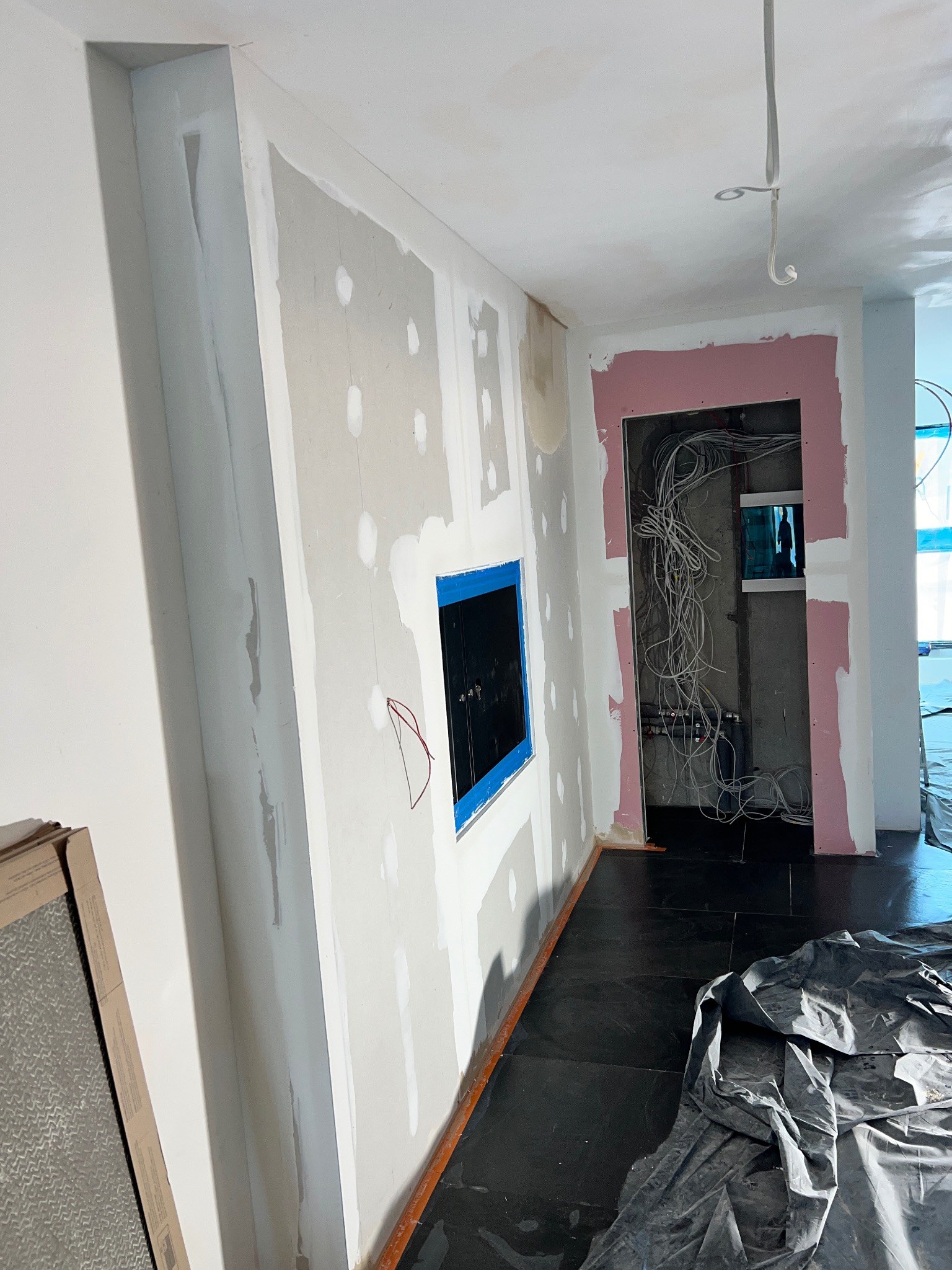 Wnętrze w trakcie remontu: ściany z nałożoną gładzią szpachlową, otwór na rozdzielnicę elektryczną zabezpieczony taśmą, widoczne przewody elektryczne w ścianie, podłoga zabezpieczona folią malarską.