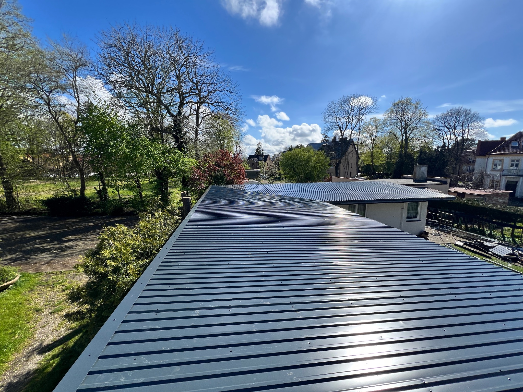 Nowy, metalowy dach w kolorze grafitowym, z widocznymi liniami i mocowaniami, błyszczący w słońcu, na tle zieleni drzew i zabudowań.