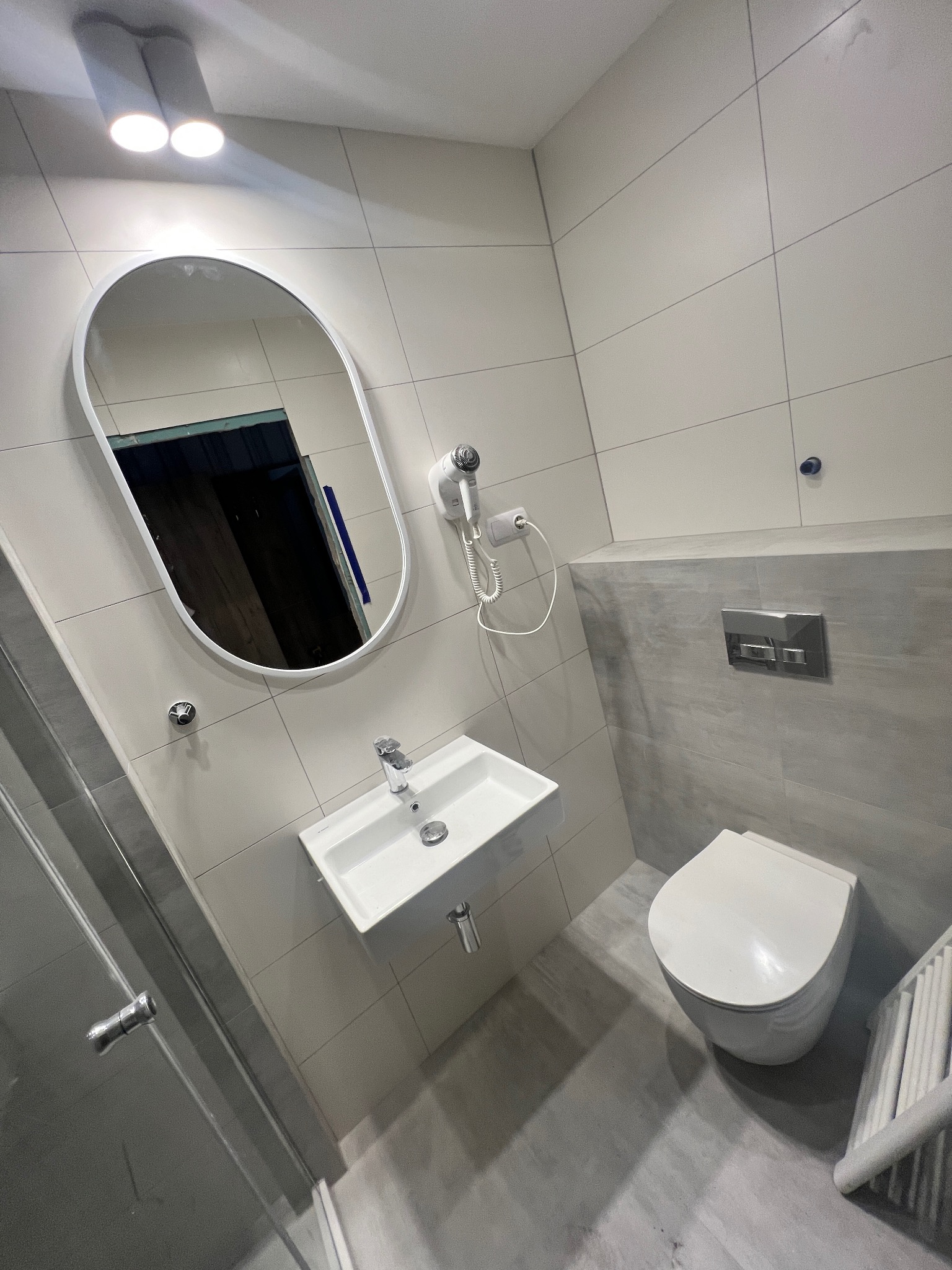 Nowoczesna łazienka z owalnym lustrem, podwieszaną umywalką, wiszącą toaletą i grzejnikiem drabinkowym, wykończona jasnymi i szarymi płytkami.