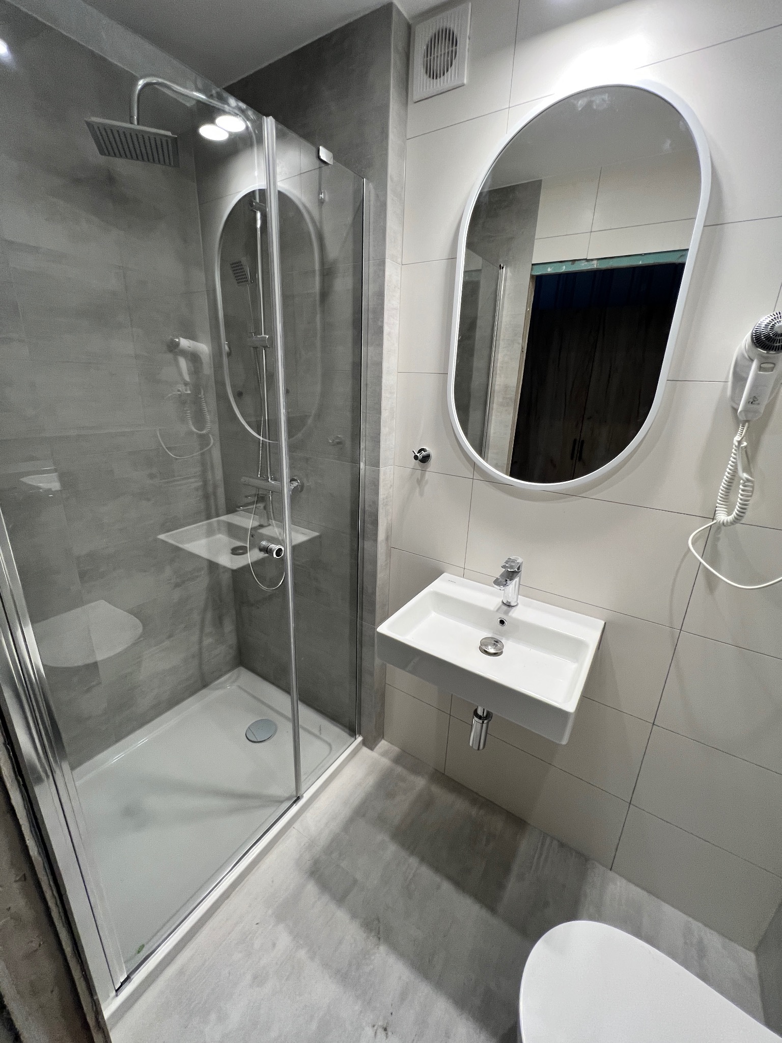 Nowoczesna łazienka z kabiną prysznicową typu walk-in, umywalką na ścianie, owalnym lustrem i suszarką do włosów. Widoczny minimalistyczny design i szara kolorystyka płytek.