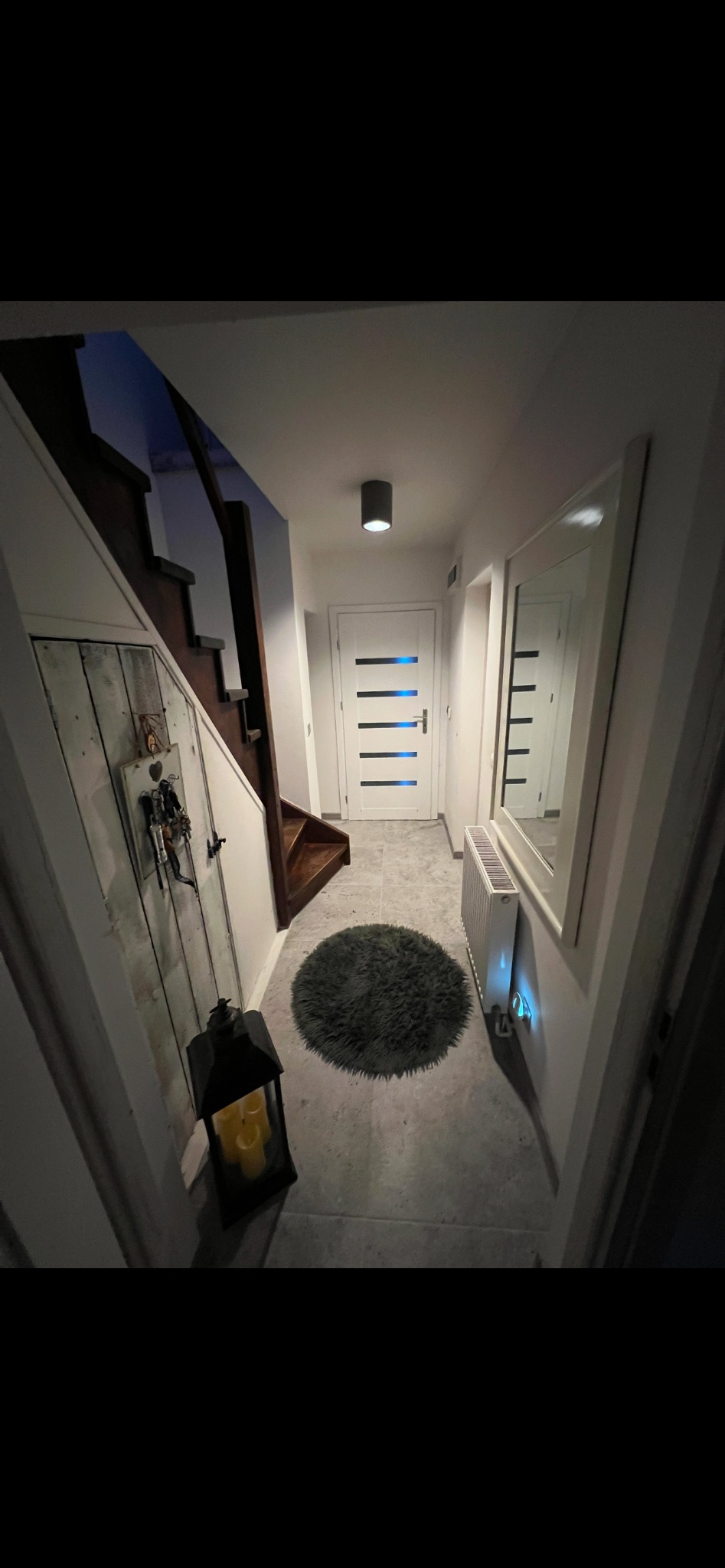 Wnętrze przedpokoju z drewnianymi schodami, szarym dywanikiem, grzejnikiem, lustrem w białej ramie i lampionem ze świecami, oświetlone punktowym światłem.