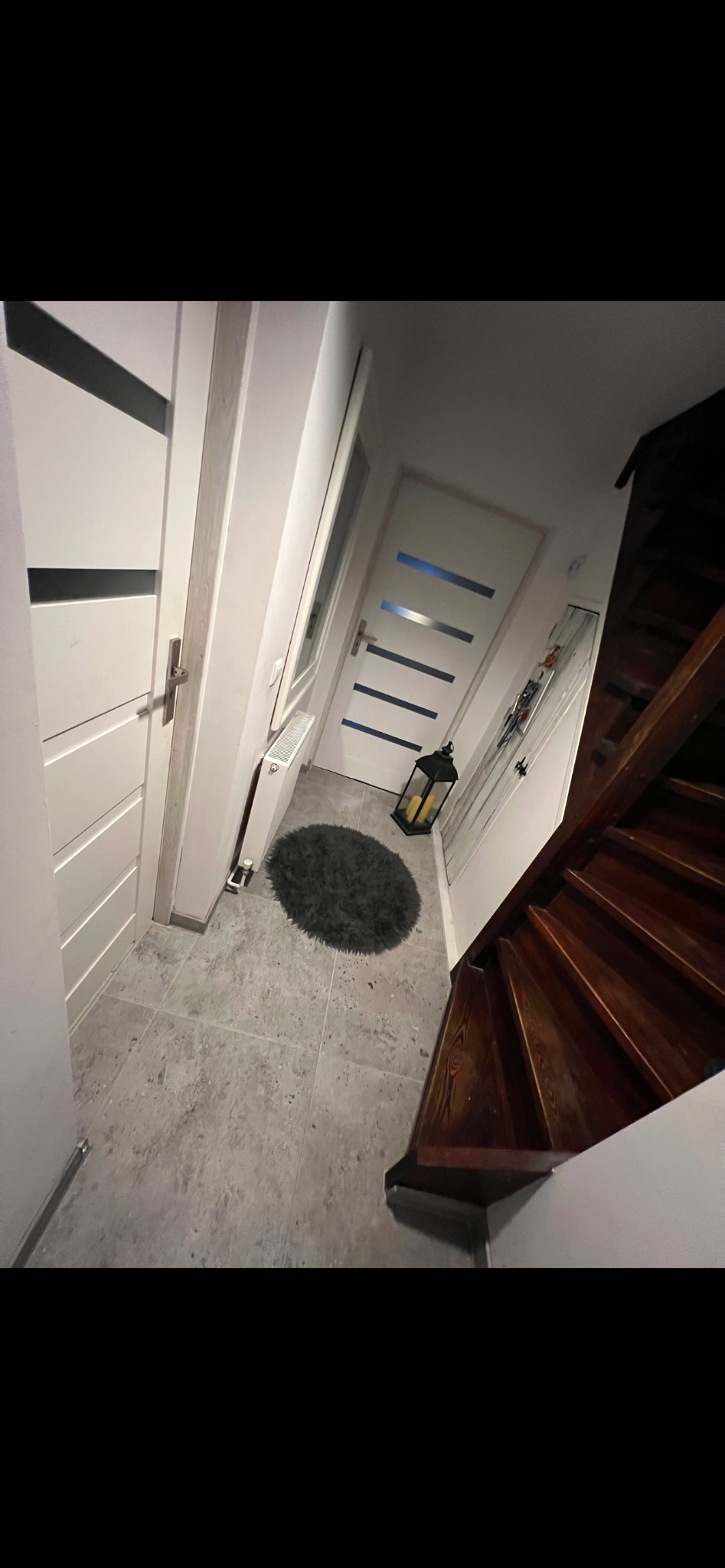 Korytarz po remoncie z szarymi płytkami podłogowymi, białymi drzwiami z czarnymi akcentami, ciemnymi drewnianymi schodami i okrągłym szarym dywanikiem.