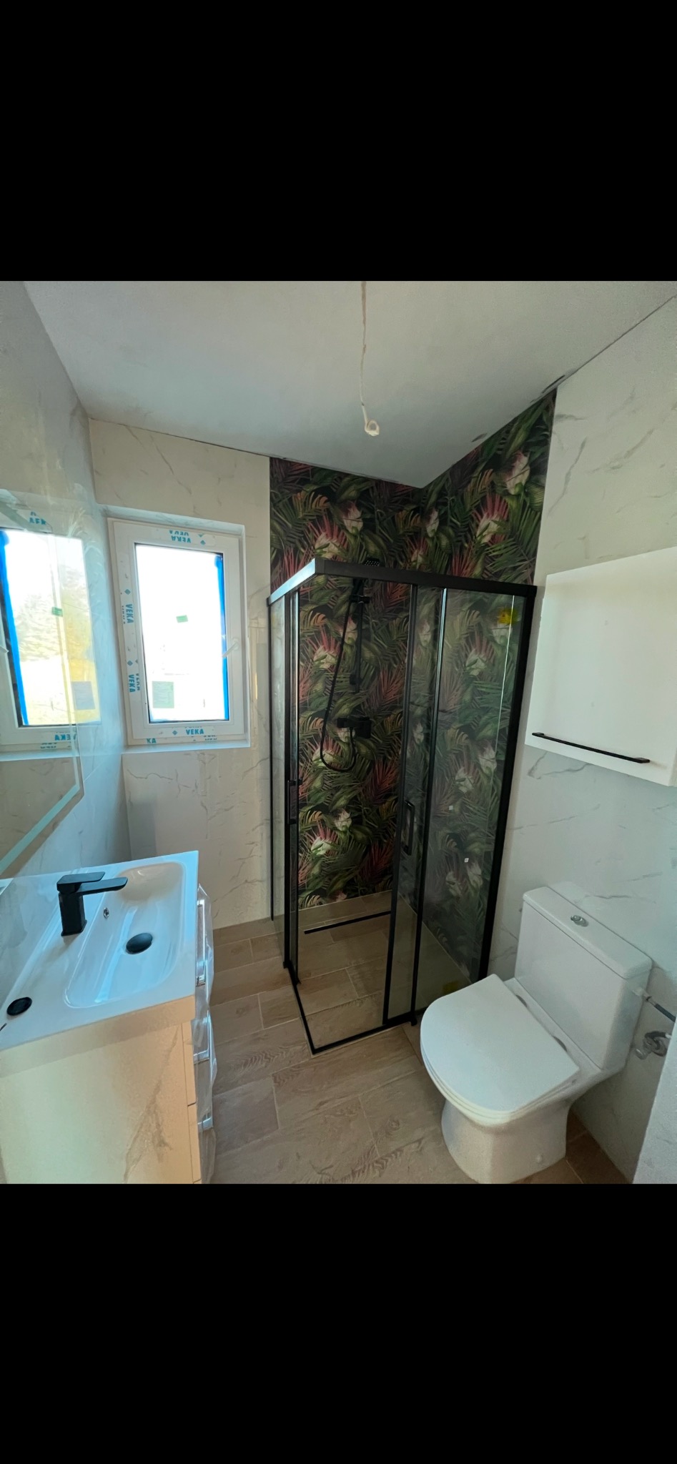 Wykończona łazienka z kabiną prysznicową z czarnymi profilami, umywalką z szafką w stylu marmuru, toaletą i tapetą w tropikalne liście na jednej ze ścian.