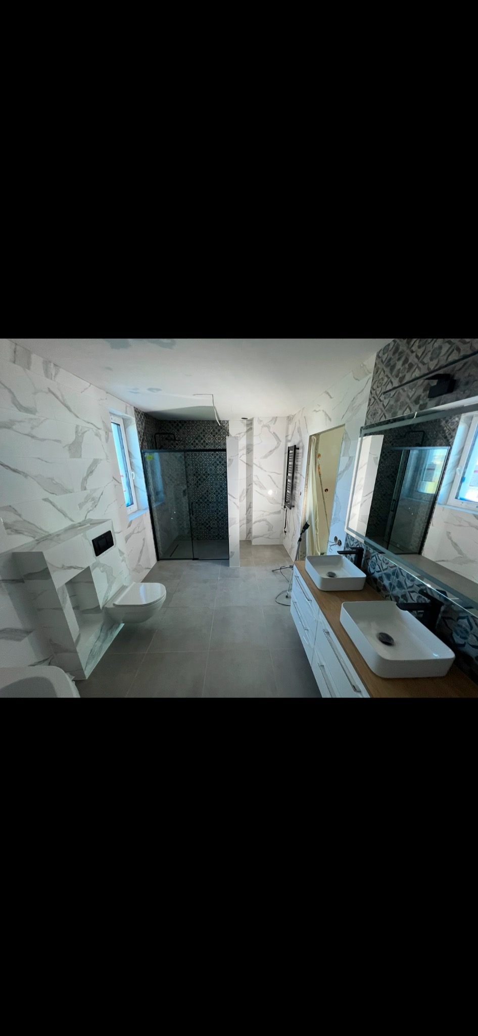 Wnętrze łazienki w trakcie remontu: białe płytki imitujące marmur na ścianach, szara podłoga, kabina prysznicowa z czarną ramą i wzorzystymi płytkami, podwieszana toaleta, dwie białe umywalki...