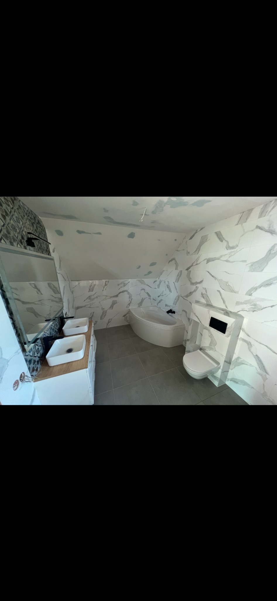 Wykończona łazienka na poddaszu z marmurowymi płytkami na ścianach, szarą podłogą, białą wanną narożną, wiszącą toaletą i podwójną umywalką na drewnianym blacie.