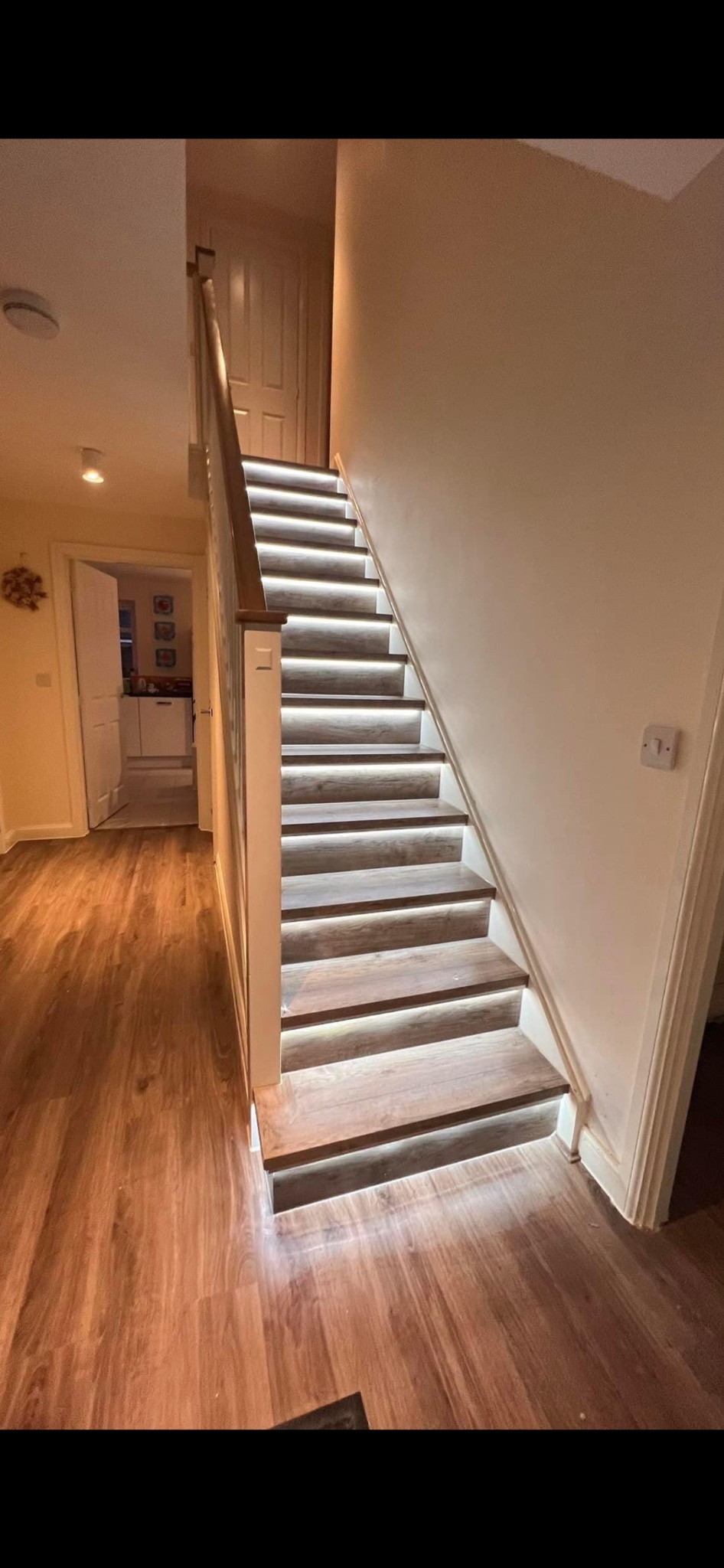 Oświetlone schody z drewnianymi stopniami i podświetleniem LED, widoczne drzwi i podłoga z paneli, ciepłe światło w pomieszczeniu.