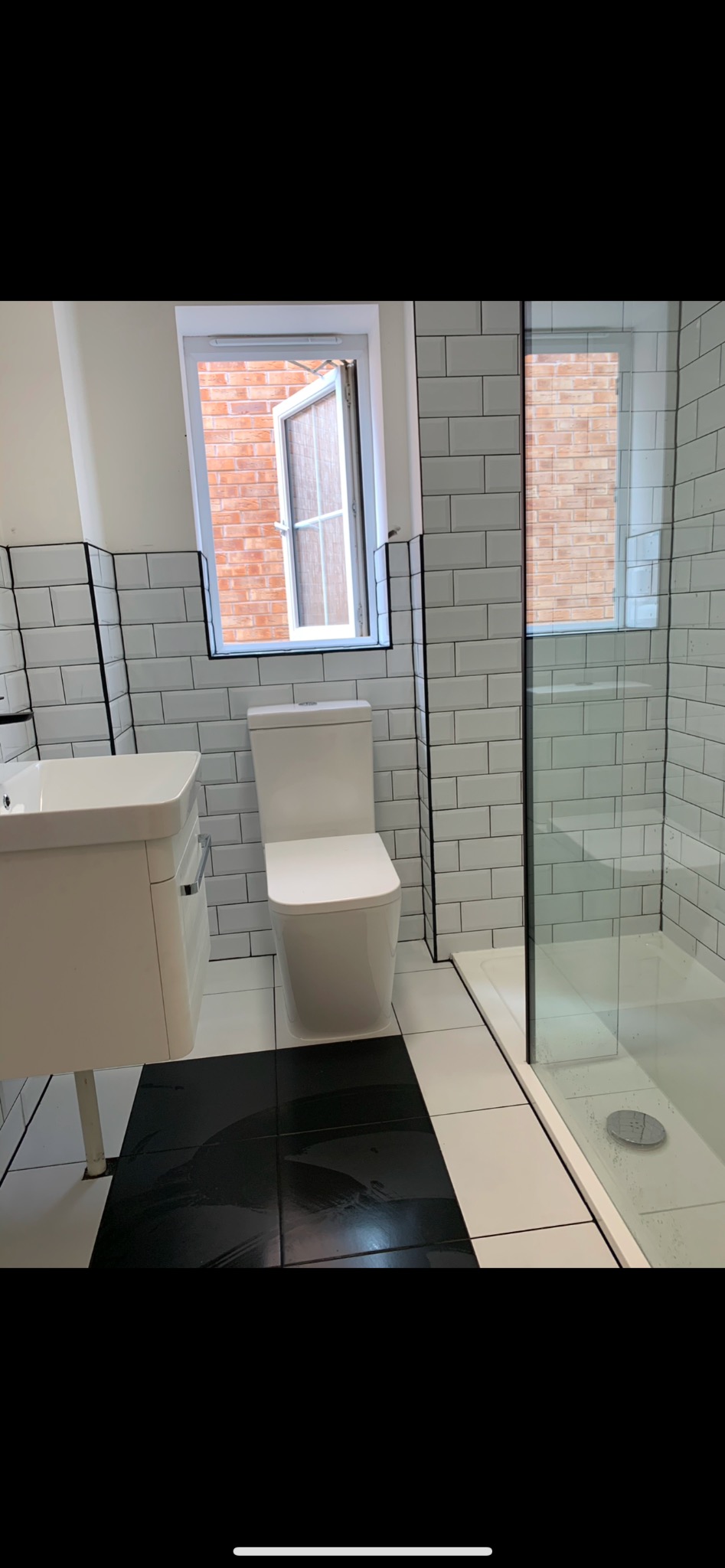 Nowoczesna łazienka po remoncie z białą armaturą, kabiną prysznicową ze szklaną ścianą, biało-czarną podłogą i białymi płytkami typu metro z czarną fugą na ścianach, widok na ceglany mur przez...