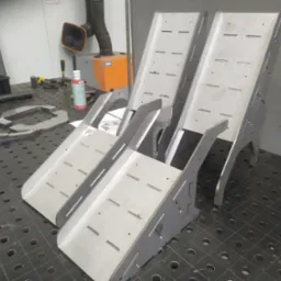 Dwie metalowe konstrukcje spawane z otworami i szczelinami, ustawione na perforowanym stole warsztatowym w warsztacie, w tle widoczne narzędzia i pochłaniacz pyłów.