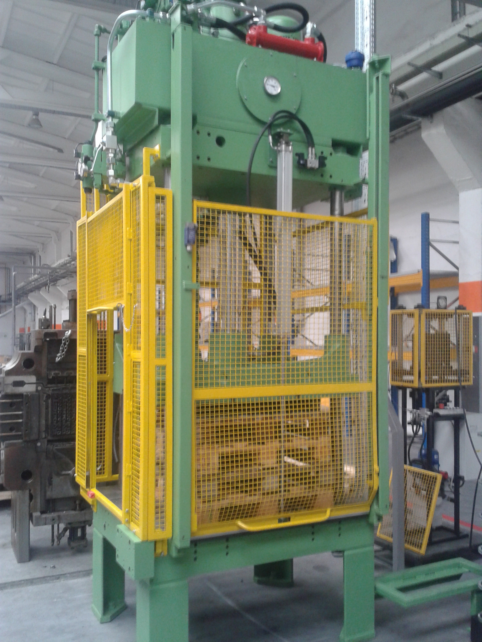 Zielona prasa hydrauliczna z żółtą siatką ochronną w hali produkcyjnej.