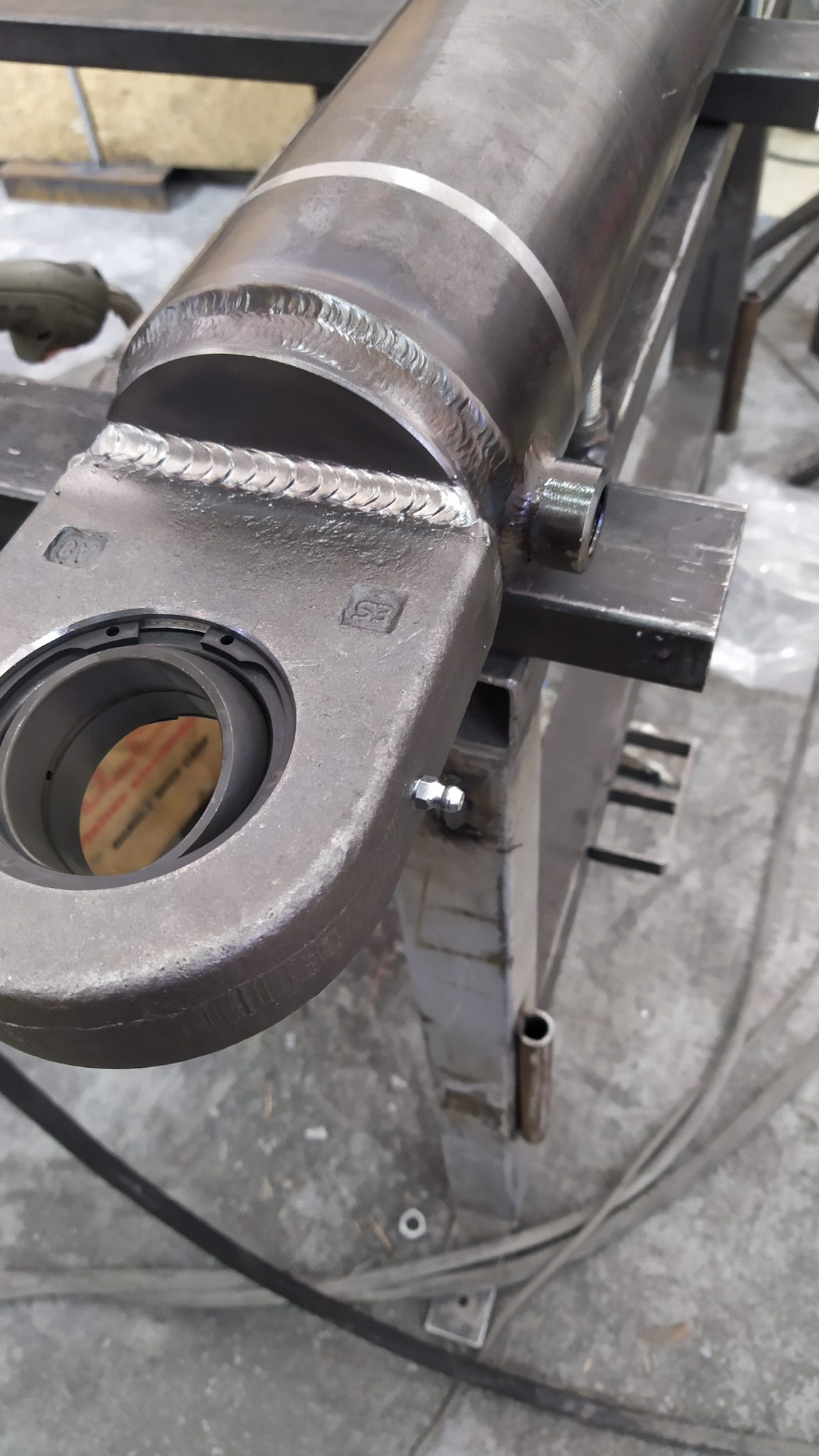 Szlifowany, spawany cylinder hydrauliczny zamontowany na metalowej konstrukcji, widoczne spawy i otwór na łożysko.