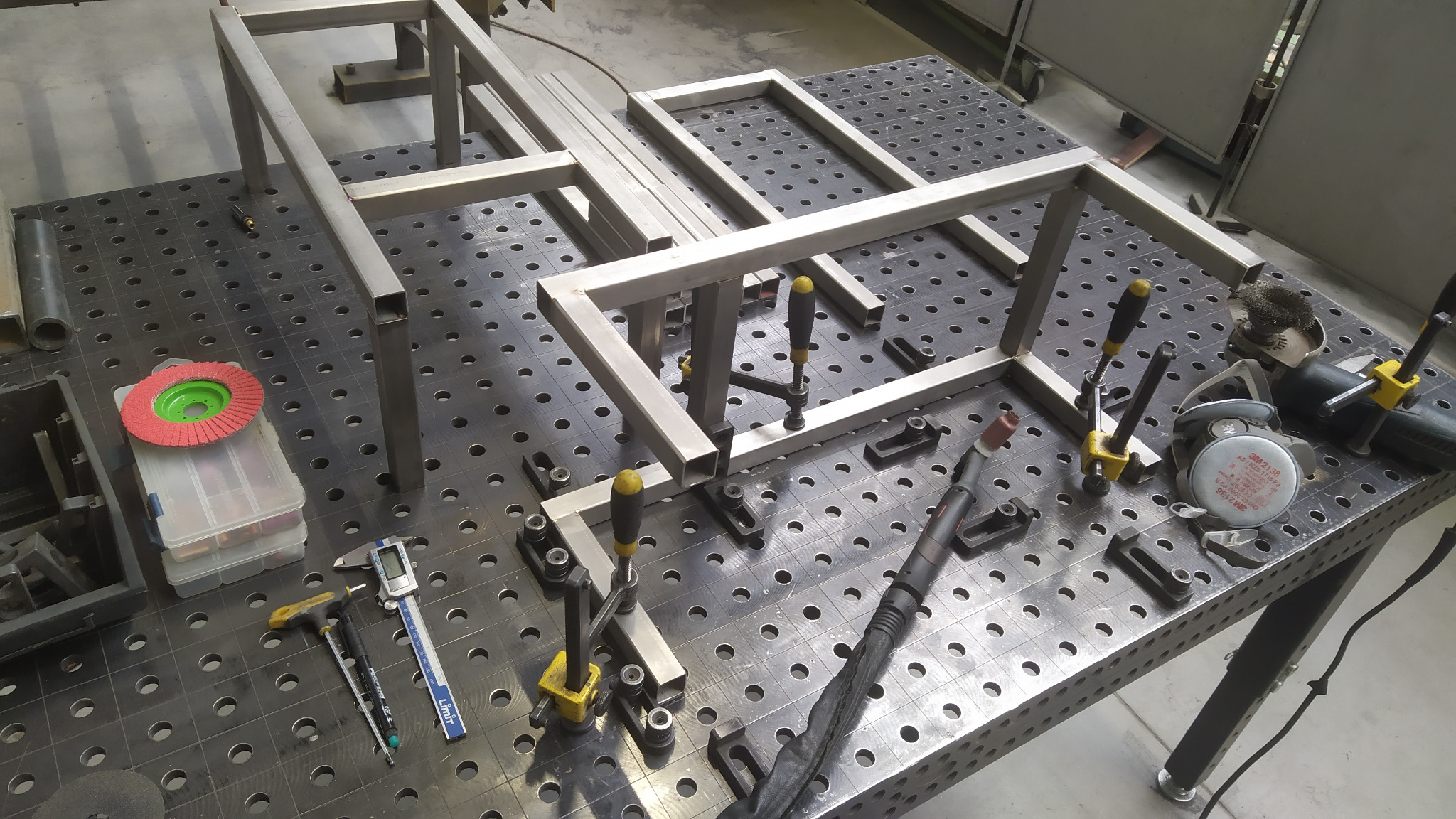 Elementy metalowe z profili kwadratowych, skręcone ściskami stolarskimi na perforowanym stole warsztatowym, obok leżą narzędzia: spawarka TIG, szlifierka kątowa, suwmiarka cyfrowa i tarcza ścierna.