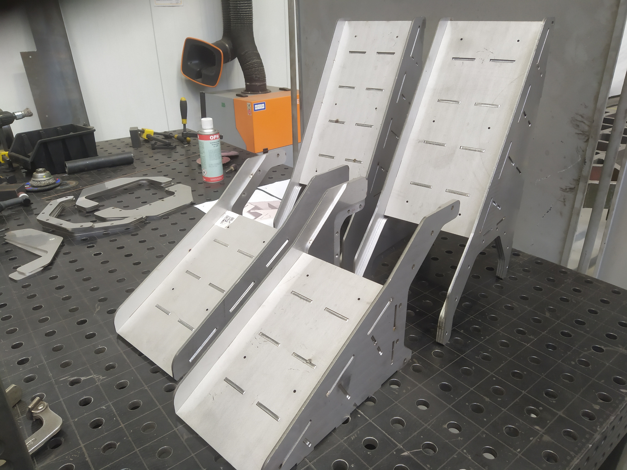 Dwie metalowe konstrukcje spawane z otworami i szczelinami, ustawione na perforowanym stole warsztatowym w warsztacie, w tle widoczne narzędzia i pochłaniacz pyłów.