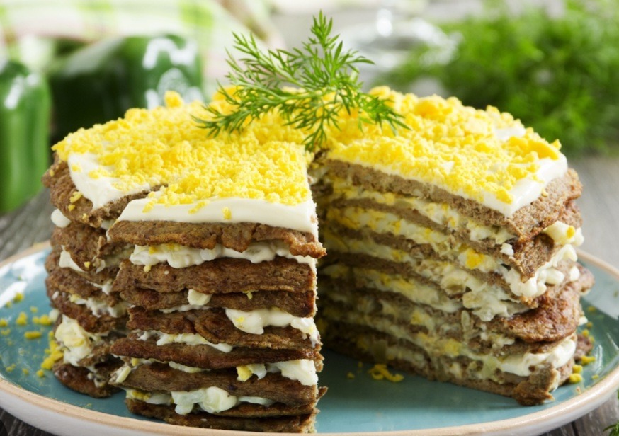 Wielowarstwowy tort z wątróbki przełożony jajkiem i majonezem, udekorowany posiekanym jajkiem na twardo i gałązką koperku, podany na turkusowym talerzu.