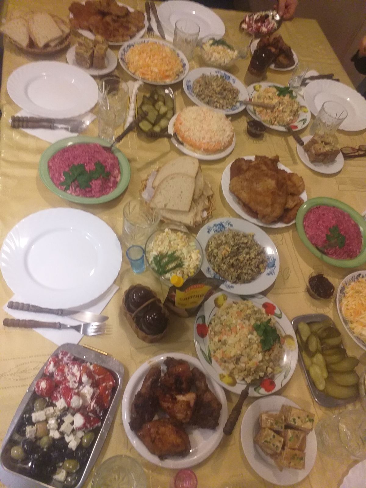 Stół zastawiony różnorodnymi potrawami, w tym sałatkami warzywnymi, mięsem smażonym, marynowanymi ogórkami, śledziem pod pierzynką, pieczywem i przekąskami, całość w ciepłej tonacji światła