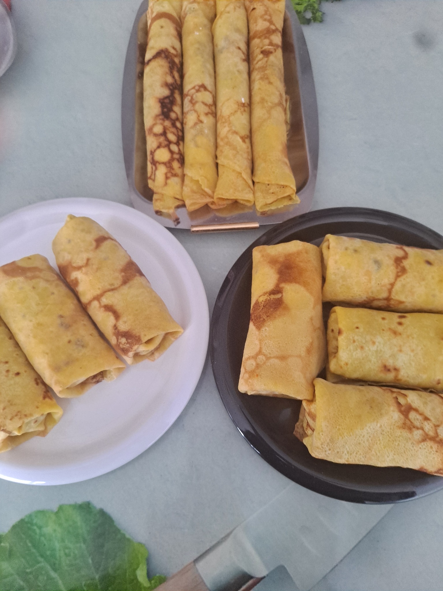 Naleśniki z farszem ułożone na trzech różnych naczyniach: srebrnej tacy, białym talerzu i czarnym talerzu, z widocznym fragmentem zielonej sałaty w dolnym rogu.