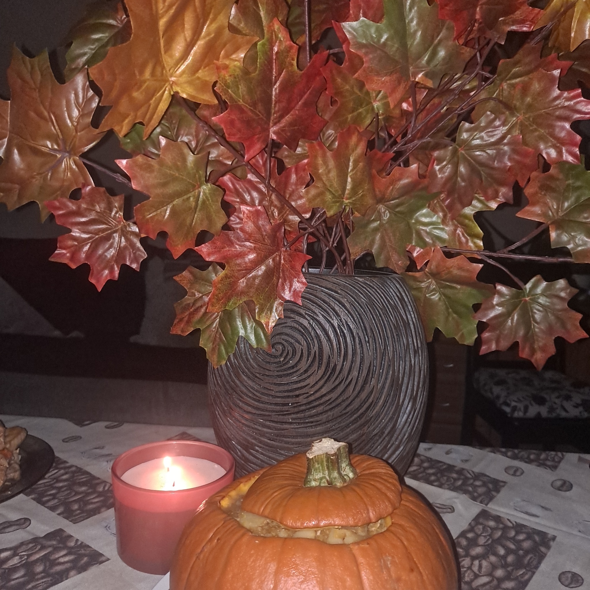 Dekoracja stołu z dynią wypełnioną potrawą, świecą w różowym szkle i wazonem z jesiennymi liśćmi na obrusie w ziarna kawy.