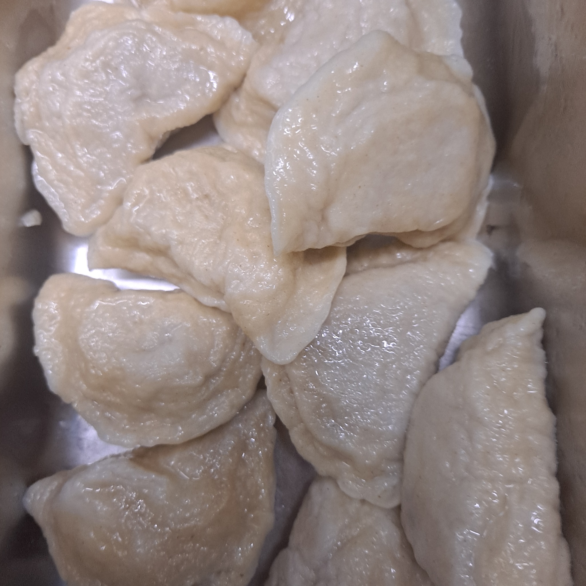 Bliski kadr gotowanych pierogów w metalowej brytfannie, oświetlonych od góry, z widoczną fakturą ciasta i delikatnym połyskiem.