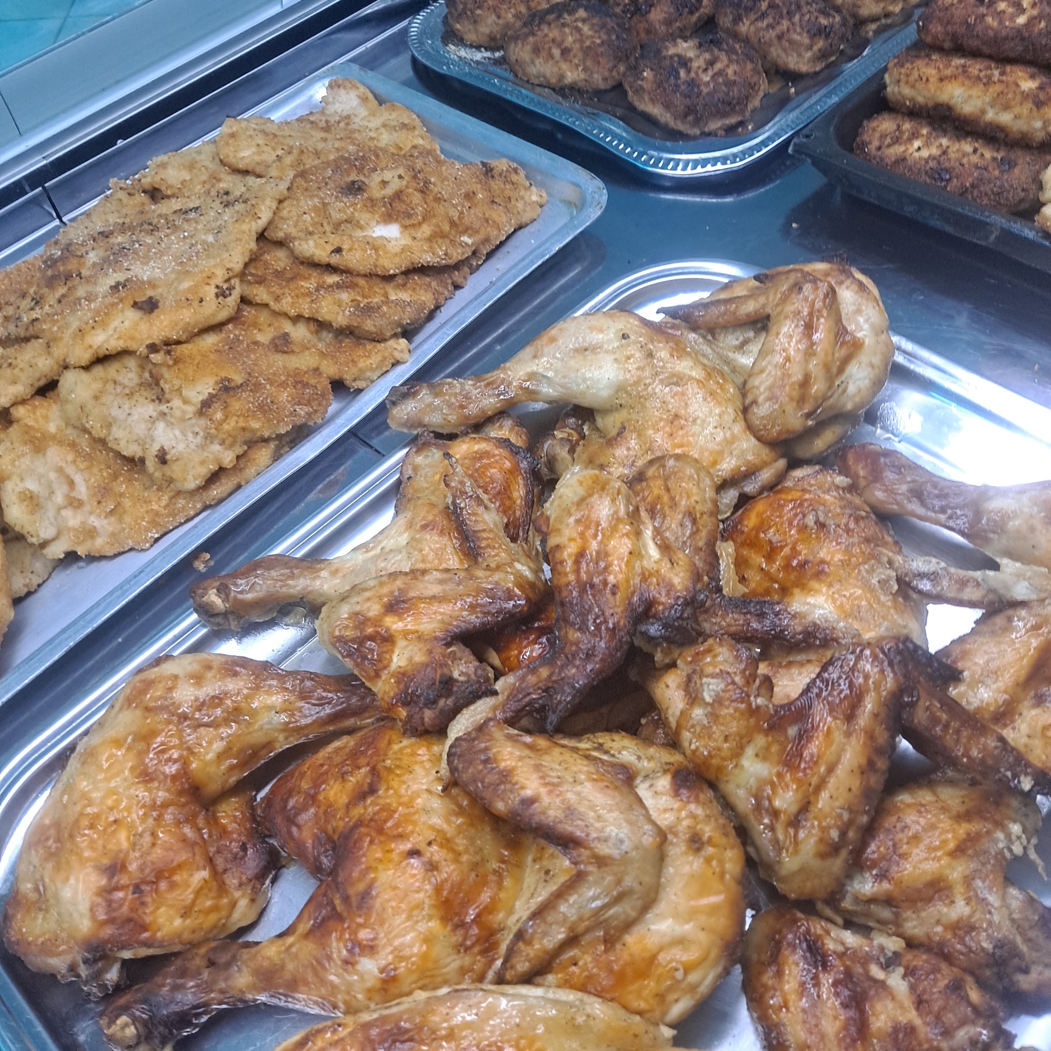 Metalowa taca z pieczonymi udkami kurczaka o złocistej skórce, obok smażone kotlety i pulpety na innych tacach, widok z góry w chłodnym oświetleniu.