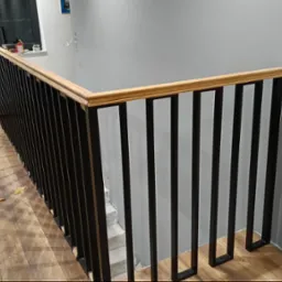 Nowoczesna balustrada schodowa z czarnymi pionowymi prętami i drewnianym poręczą, widoczna z góry, z elementami wykończeniowymi na podłodze.