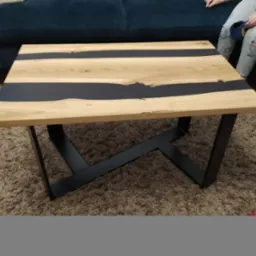 Stolik kawowy z blatem z jasnego drewna z żywicą epoksydową w kolorze czarnym, na czarnych, metalowych nogach w kształcie litery X, na tle pluszowego dywanu i ciemnozielonej sofy.