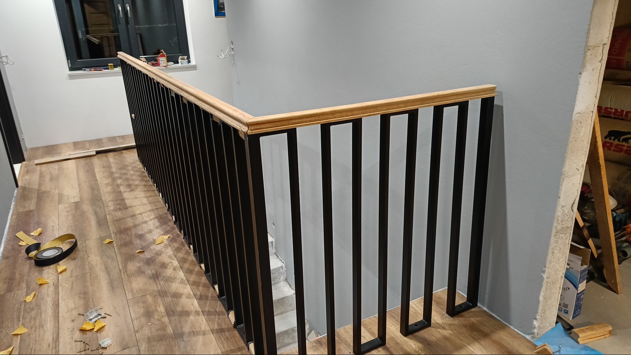 Nowoczesna balustrada schodowa z czarnymi pionowymi prętami i drewnianym poręczą, widoczna z góry, z elementami wykończeniowymi na podłodze.