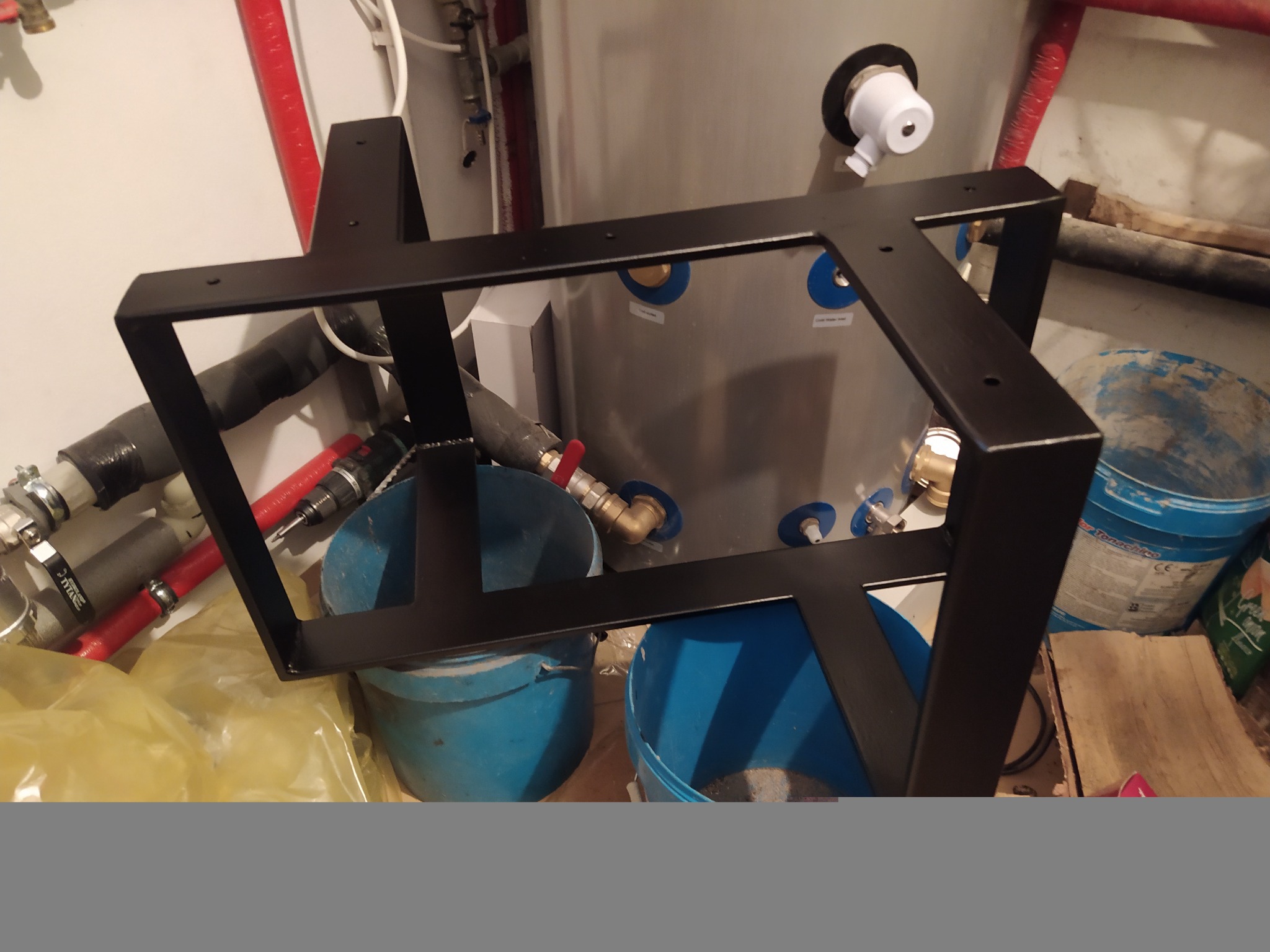 Czarna, stalowa konstrukcja pod rekuperator, widoczne niebieskie wiadra i instalacja hydrauliczna w tle.