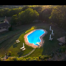 Malisz Film Studio - Panoramiczny widok z lotu ptaka na urokliwy basen o nieregularnym kształcie, otoczony zielonym trawnikiem, leżakami i parasolami, w scenerii toskańskiej willi z czerwonym dachem na wzgórzu...