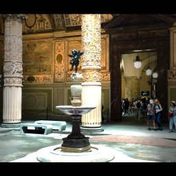 Malisz Film Studio - Wnętrze renesansowego pałacu z kolumnami zdobionymi płaskorzeźbami, fontanną pośrodku i turystami w tle, oświetlone naturalnym światłem wpadającym przez drzwi.